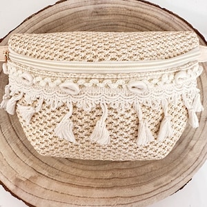 Op de afbeelding: Een crèmekleurige geweven strooien heuptas met een ritssluiting en een witte kwastversiering. De tas heeft een witte gehaakte rand met schelpvormige accenten.