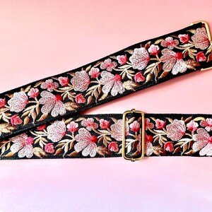 Könnte beinhalten: Schwarzer, bestickter Taschenriemen mit einem floralen Design in Rosa, Weiß und Gold. Der Riemen hat goldfarbene Metallbeschläge, einschließlich Verschlüssen und einer verstellbaren Schnalle. Der Riemen ist vor einem rosa Hintergrund platziert.