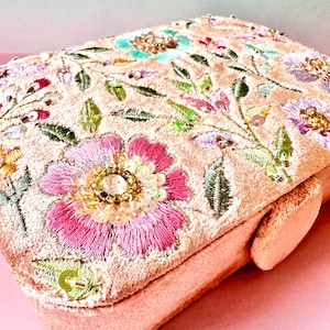 Könnte beinhalten: Eine pfirsichfarbene, rechteckige Clutch mit aufwendiger Blumenstickerei. Die Blumen sind rosa, türkis und lila, verziert mit Pailletten und Perlen. Die Clutch hat einen passenden Pfirsichverschluss und eine weiche, strukturierte Optik.