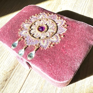 Pode incluir: Uma caixa de joias retangular de veludo rosa empoeirado. A tampa é adornada com um desenho floral bordado em ouro, roxo e creme, centrado com uma gema roxa. Três pingentes de cristal em forma de gota pendem do desenho.