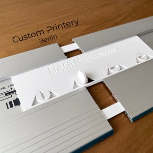 Può includere: Uno strumento bianco e argento con la scritta "BOSCH FSN Verbinder" e "Custom Printery Berlin". Lo strumento sembra essere un connettore per un sistema di guide Bosch FSN. L'immagine mostra lo strumento parzialmente assemblato.