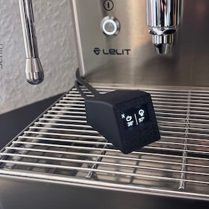 Op de afbeelding: Een zwart, rechthoekig apparaat met een klein digitaal display met temperatuurmetingen, geplaatst op een roestvrijstalen espressomachine. De machine heeft een Lelit-logo en een chromen stoompijp. De tekst "Custom Printery Berlin" is zichtbaar.