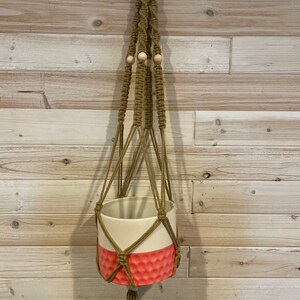 May include: A macrame plant hanger with a white and pink ceramic planter. The hanger is made of brown cord and has wooden beads.