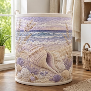 Könnte beinhalten: Ein zylindrischer Wäschekorb aus Stoff mit einer gesteppten Küstenszene, die eine große Muschel, verschiedene kleinere Muscheln, Sanddollars und hohes Strandgras in Lavendel-, Beige- und Blautönen zeigt, mit zwei Tragegriffen.