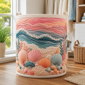 May include: A cylindrical laundry hamper featuring a quilted design of an ocean scene with pink and orange sunset waves, teal water, and a sandy beach foreground decorated with embroidered seashells in shades of pink, orange, and blue.