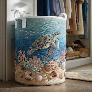 May include: A cylindrical laundry hamper featuring a quilted ocean scene with a sea turtle swimming above a sandy seabed. The design includes coral, various seashells, and small fish in shades of blue, tan, and pink, with two white fabric handles.