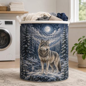 May include: A cylindrical laundry hamper featuring a detailed print of a grey wolf standing in a snowy forest landscape at night under a full moon. The hamper has a white top rim and two white side handles, filled with folded fabric items.