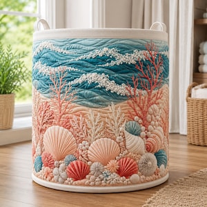 May include: A cylindrical fabric laundry hamper featuring a quilted design of ocean waves in shades of blue and white above a sandy beach scene decorated with textured, three-dimensional coral and various seashells in pink, peach, and blue.