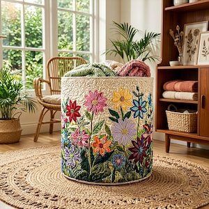 May include: A cylindrical fabric storage basket featuring a textured cream-colored base with a vibrant, multi-colored floral pattern. The exterior displays embroidered flowers in shades of pink, yellow, blue, orange, and purple with green stems.