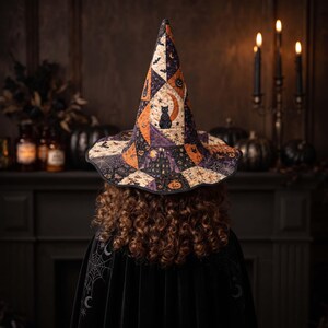 May include: A tall, pointed witch hat featuring a patchwork design with various Halloween-themed patterns including black cats, crescent moons, pumpkins, and haunted houses in shades of orange, purple, black, and cream, worn over curly hair.