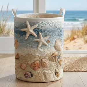 Könnte beinhalten: Ein zylindrischer Wäschekorb aus Stoff mit gestepptem Design im Strand-Thema. Die Außenseite zeigt ein Muster aus blauen Meereswellen, sandfarbenen Küsten sowie verschiedenen Muscheln und Seesternen, die auf der Oberfläche appliziert sind.