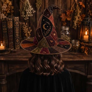 Puede incluir: Un sombrero puntiagudo con un diseño de parches de terciopelo y tela en tonos burdeos, verde oliva y negro. El sombrero está decorado con motivos celestiales bordados, que incluyen una luna creciente, estrellas y elementos botánicos.