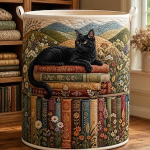 May include: A cylindrical fabric laundry hamper featuring a quilted design of a black cat resting on a stack of vintage books. The exterior displays a landscape of rolling hills, wildflowers, and a row of colourful book spines with floral patterns.