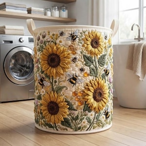 May include: A cylindrical fabric laundry basket featuring a quilted texture with an embroidered pattern of large yellow sunflowers, green leaves, small white flowers, and several bees. The design includes a honeycomb motif with amber accents.