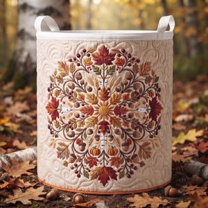 May include: A cylindrical fabric storage basket with a cream-colored quilted exterior featuring a central embroidered mandala design of autumn leaves, acorns, berries, and small pumpkins. The basket has two white fabric handles at the top rim.