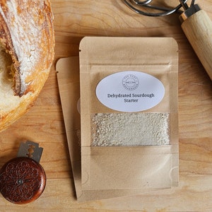 Peut inclure: Un sachet en papier brun avec une fenêtre transparente montrant un levain de pain au levain déshydraté. Le sachet porte une étiquette ovale blanche avec le texte "The Busy Breadmaker" et "Dehydrated Sourdough Starter".