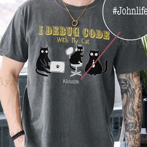 Peut inclure: T-shirt gris foncé avec un motif de trois chats noirs dessinés. Le texte "I DEBUG CODE With My Cat" est en or, avec le hashtag "#Johnlife" en dessous. Le t-shirt a un aspect vintage et usé.