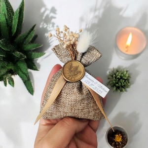 以下が含まれることがあります： 金色のワックスシールと「Amanda & Matthew 05.17.2025」と書かれた白いタグが付いた小さな麻袋。袋はドライフラワーで満たされ、金色のリボンで結ばれています。