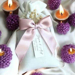 Puede incluir: Una bolsa de lino blanca con una cinta de satén rosa y una pequeña etiqueta que dice "Gizem & Arif Nisan Hatırası 27.07.2024". La bolsa está rodeada de velas con forma de flores moradas.
