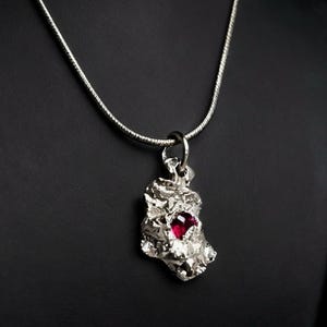 May include: A silver pendant necklace. The pendant has a textured, irregular shape with a red gemstone. The necklace hangs from a delicate silver chain. The background is dark.