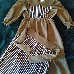 May include: Olive green dress with long sleeves and a Peter Pan collar. The bodice features a vertical black and white striped pattern. The dress has a gathered waist and a flowing skirt. The sleeves have ruffled cuffs.
