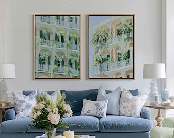 May include: Two framed rectangular wall art prints depicting multi-story buildings with balconies and lush green foliage, displayed side-by-side above a blue sofa. The paintings feature soft pastel tones, architectural details, and hanging plants.