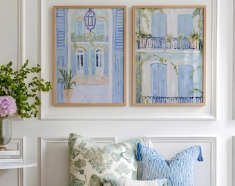 May include: Two framed paintings of buildings with blue shutters hang above a bench with green cushions and decorative pillows. A small side table with a gold base and a vase of flowers is to the left.