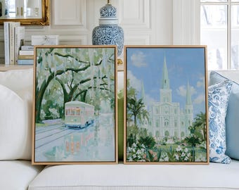 May include: Two rectangular framed wall art prints featuring New Orleans scenes. The left print depicts a green streetcar moving through a tree-lined street with hanging moss. The right print shows a white cathedral with spires and lush gardens.