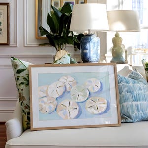 May include: A framed artwork featuring a watercolor painting of sand dollars in shades of white, beige, and blue. The artwork is displayed on a white sofa, with decorative pillows in shades of blue and green. A blue and white ceramic lamp and a gold-framed mirror are also visible.