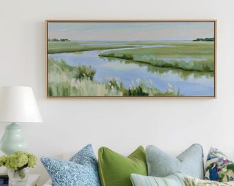 May include: A rectangular landscape painting featuring a marshland with a winding blue river, green grasses, and a soft sky, framed in light wood. The artwork hangs above a white sofa adorned with various blue, green, and patterned throw pillows.