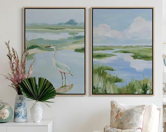 May include: Two framed landscape paintings featuring a heron in a marshland setting with blue water, green grasses, and a cloudy sky. The artwork is displayed above a white cabinet with a vase of dried flowers and a white armchair with patterned pillows.