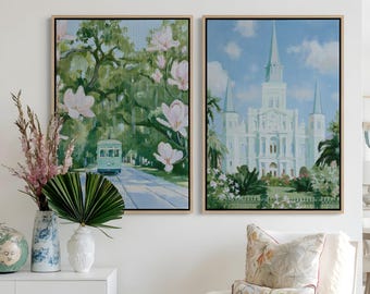May include: Two framed rectangular wall art prints featuring New Orleans scenes. The left print shows a green streetcar under oak trees with pink magnolia blossoms. The right print depicts a white cathedral against a blue sky with clouds.