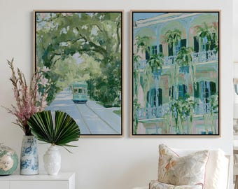May include: Two framed rectangular wall art prints featuring impressionistic scenes. The left print depicts a green streetcar on a tree-lined road. The right print shows a multi-story building with ornate balconies, green shutters, and hanging plants.