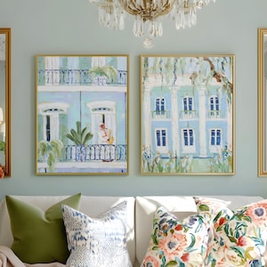 May include: Two framed paintings of buildings with blue and white facades hang above a white sofa with decorative pillows. The paintings have gold frames. The sofa has green, blue, and floral pillows.
