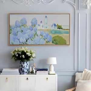 Op de afbeelding: Een rechthoekig landschapsschilderij in een lichthouten lijst met een vuurtoren en gebouwen bij een veld met blauwe hortensia's. Het kunstwerk toont een kusttafereel met zachte blauwe, groene en beige tinten en legt een sereen uitzicht vast.