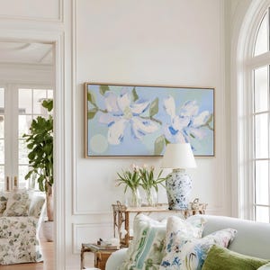 May include: A rectangular wall art piece featuring a painted floral design with large white and blue blossoms on a light blue background, framed in a light wood border, displayed above a sofa with various patterned and solid green throw pillows.