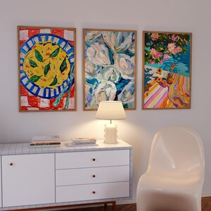K&ouml;nnte beinhalten: Drei gerahmte Bilder an einer wei&szlig;en Wand &uuml;ber einer wei&szlig;en Kommode mit drei Schubladen. Die Bilder zeigen einen Teller Pasta, einen Strau&szlig; wei&szlig;er Blumen und eine Strandszene mit einem gestreiften Handtuch und Obst.