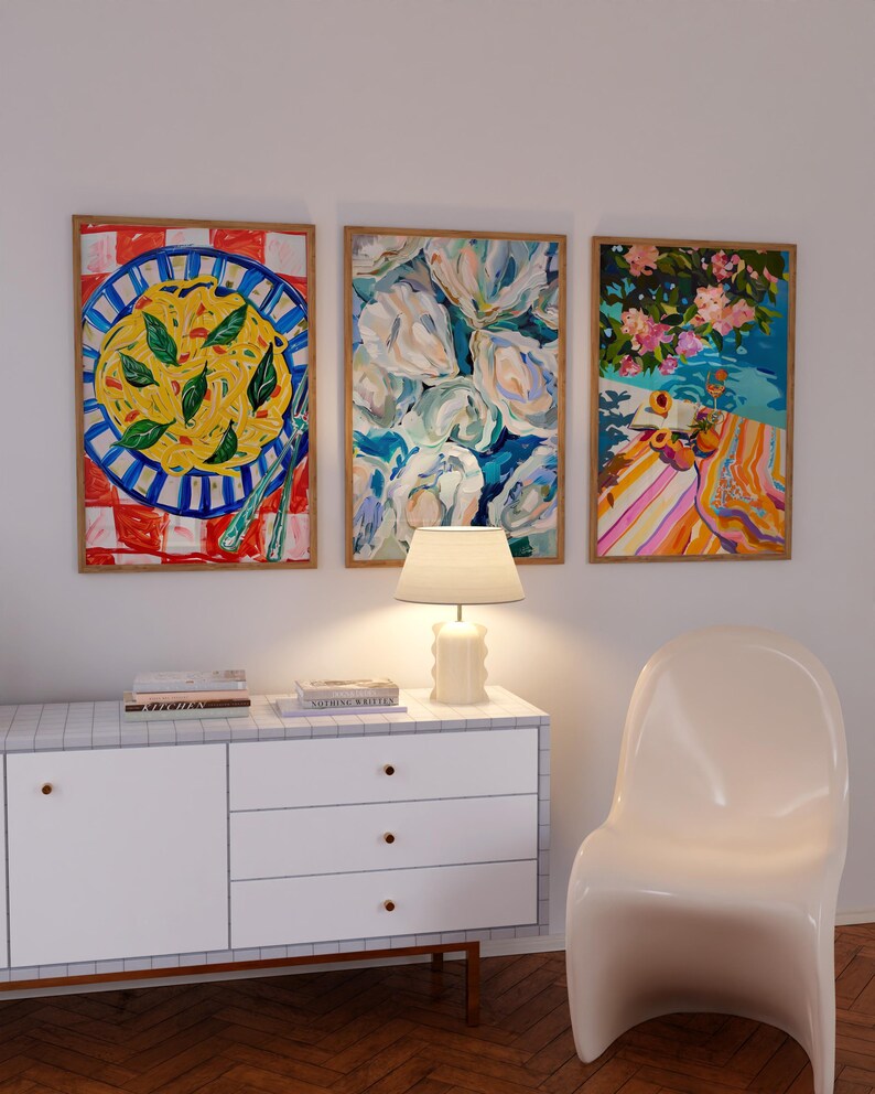 K&ouml;nnte beinhalten: Drei gerahmte Bilder an einer wei&szlig;en Wand &uuml;ber einer wei&szlig;en Kommode mit drei Schubladen. Die Bilder zeigen einen Teller Pasta, einen Strau&szlig; wei&szlig;er Blumen und eine Strandszene mit einem gestreiften Handtuch und Obst.