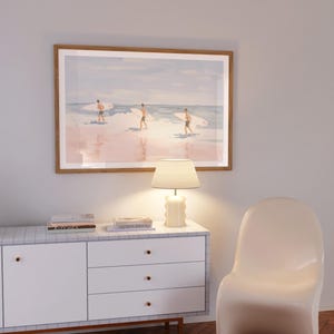 May include: A white dresser with three drawers and a light wood top. A lamp with a white shade sits on the top of the dresser. A framed print of three surfers on a beach hangs on the wall above the dresser. A white chair sits in the corner of the room.