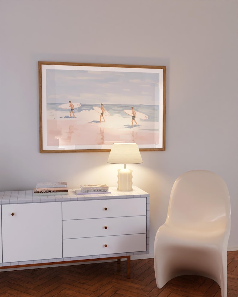 May include: A white dresser with three drawers and a light wood top. A lamp with a white shade sits on the top of the dresser. A framed print of three surfers on a beach hangs on the wall above the dresser. A white chair sits in the corner of the room.