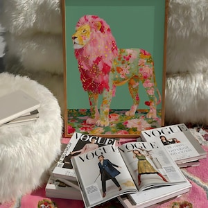 K&ouml;nnte beinhalten: Gerahmter Kunstdruck eines L&ouml;wen mit rosa M&auml;hne und floralem K&ouml;rper. Der L&ouml;we steht auf einer floral gemusterten Oberfl&auml;che vor einem gr&uuml;nen Hintergrund. Vogue-Magazine sind vor dem Druck gestapelt.