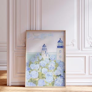 May include: Framed art print featuring two white lighthouses with blue accents, set against a backdrop of blue hydrangea flowers. The artwork is in a gold frame and is displayed in a room with white paneling and a wooden floor.