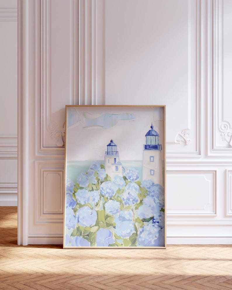 May include: Framed art print featuring two white lighthouses with blue accents, set against a backdrop of blue hydrangea flowers. The artwork is in a gold frame and is displayed in a room with white paneling and a wooden floor.