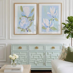 May include: Two framed floral art prints with light blue and white flowers hang above a mint green cabinet with gold hardware. A white sofa and a wooden coffee table with books are in the foreground. A vase of white roses sits on the table.