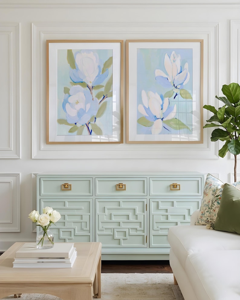 May include: Two framed floral art prints with light blue and white flowers hang above a mint green cabinet with gold hardware. A white sofa and a wooden coffee table with books are in the foreground. A vase of white roses sits on the table.