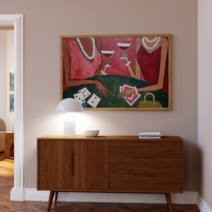 May include: A wooden mid-century modern style cabinet with three drawers and two doors. Above the cabinet is a framed painting of two figures holding glasses of red wine. A white lamp and bowl sit on top of the cabinet.