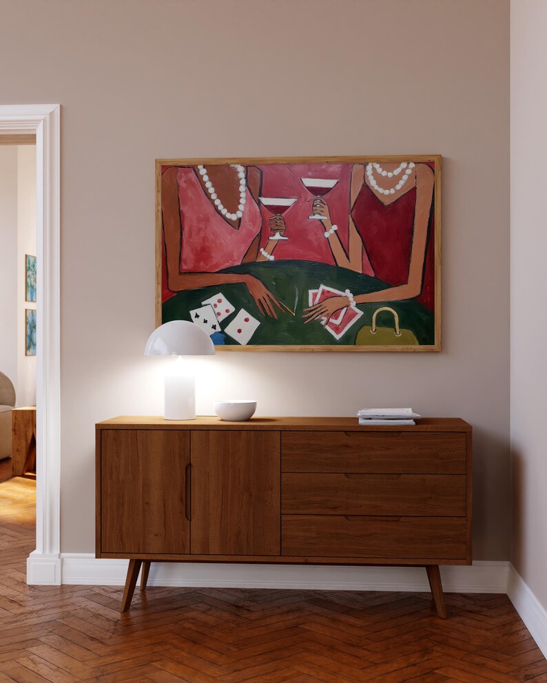 May include: A wooden mid-century modern style cabinet with three drawers and two doors. Above the cabinet is a framed painting of two figures holding glasses of red wine. A white lamp and bowl sit on top of the cabinet.