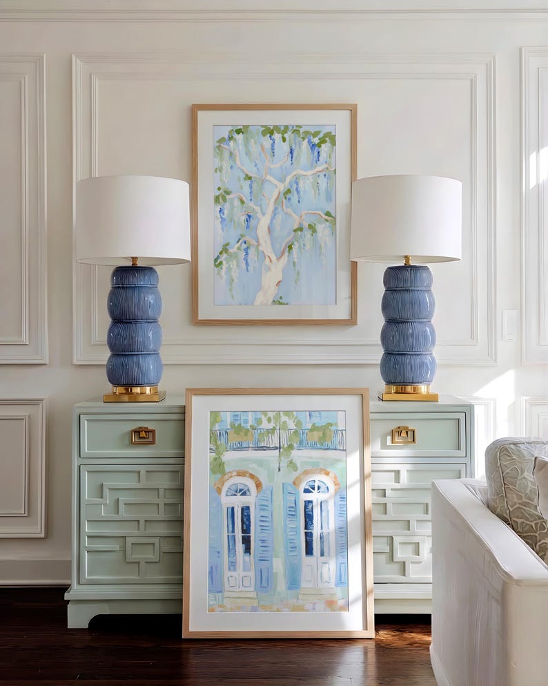 Puede incluir: Dos l&aacute;mparas de cer&aacute;mica azul con pantallas blancas se asientan sobre gabinetes azul claro a juego. Una pintura en acuarela enmarcada de un &aacute;rbol cuelga entre las l&aacute;mparas. Otra pintura enmarcada de un edificio con contraventanas azules est&aacute; al frente.