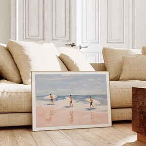 May include: A framed watercolor painting of three people walking on a beach with surfboards. The beach is pink and the water is blue. The sky is light blue with white clouds.