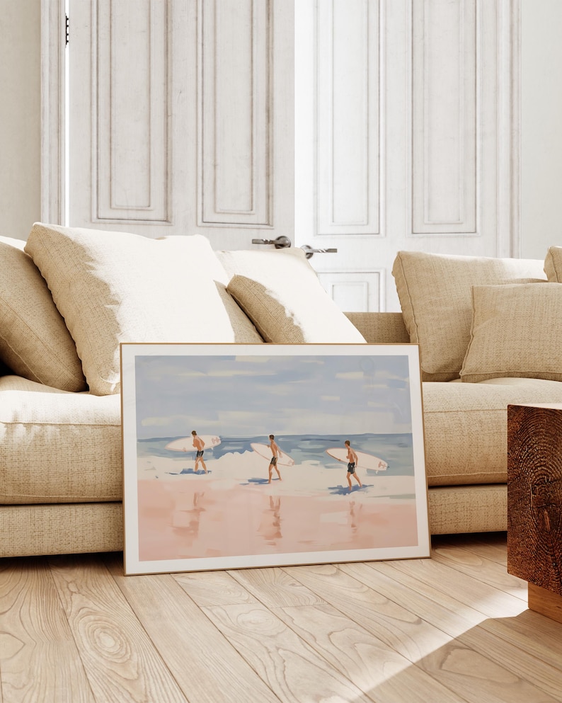 May include: A framed watercolor painting of three people walking on a beach with surfboards. The beach is pink and the water is blue. The sky is light blue with white clouds.